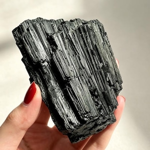 May include: A raw black tourmaline crystal specimen featuring a deeply striated, columnar structure with a glossy, reflective surface. The mineral displays a jagged, irregular top edge and a dense, opaque black coloration throughout its form.