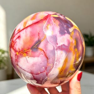 Peut inclure: Une pierre sphérique polie présentant un motif marbré vibrant avec des tourbillons de rose vif, de violet profond, d'orange chaud et de jaune. La surface affiche des inclusions complexes et fines en forme de veines rouge foncé et noires.