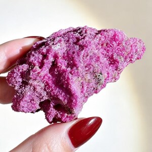 May include: A raw, crystalline mineral specimen with a vibrant, saturated magenta-pink hue. The stone features a highly textured, jagged surface composed of numerous small, sparkling crystal facets that catch the light across its irregular shape.