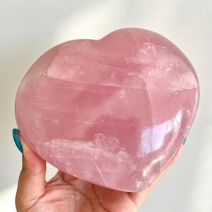 May include: A polished heart-shaped rose quartz crystal with a translucent, pale pink hue. The stone features natural internal fractures and variations in color, showcasing a smooth, glossy surface texture throughout the entire piece.