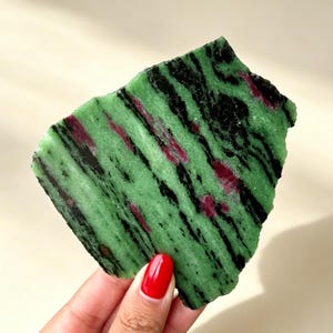 Puede incluir: Una losa plana de mineral de zoisita de forma irregular que presenta una base de color verde vibrante con prominentes vetas negras alargadas e inclusiones de color rojo magenta intenso dispersas por la superficie pulida.