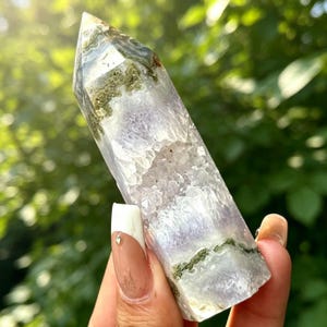 Puede incluir: Una torre de cristal hexagonal pulida que presenta una base de amatista translúcida de color púrpura pálido con estructuras cristalinas internas e inclusiones minerales de color verde musgo concentradas cerca de la punta y la base.
