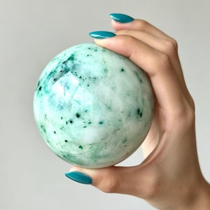 Puede incluir: Una piedra esférica pulida con una superficie lisa y brillante. La piedra presenta un color base verde menta pálido con intrincadas inclusiones minerales de color verde oscuro y motas negras dispersas por toda su forma redondeada.