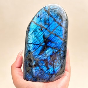 May include: A polished labradorite stone featuring a vibrant blue iridescent flash across its surface. The stone has a smooth, rounded top and displays dark, intersecting mineral veins that create a complex, fractured pattern throughout.