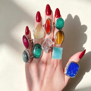May include: A collection of ten silver rings featuring various gemstones including a blue lapis lazuli, a green malachite, a clear quartz, a tiger's eye, a red carnelian, a pink stone, a yellow rutilated quartz, and a light blue stone.