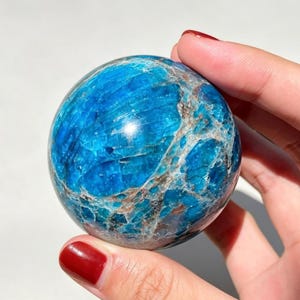 May include: A polished spherical blue apatite gemstone featuring a vibrant cyan and deep blue color palette with intricate, naturally occurring white and brown mineral veining throughout the smooth, rounded surface of the stone.