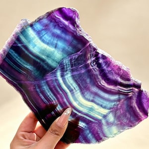 Pode incluir: Uma fatia polida de mineral fluorite com faixas vibrantes de roxo profundo, violeta e azul-petróleo translúcido. A pedra apresenta uma forma natural e irregular com uma superfície lisa e brilhante que destaca o zoneamento de cores distinto e as camadas cristalinas internas em todo o espécime.