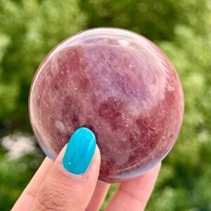 Peut inclure: Une sphère lisse et polie composée d'un minéral rose foncé translucide. La pierre présente des variations internes naturelles, des inclusions blanches nuageuses et une surface brillante qui reflète la lumière sur sa forme arrondie.
