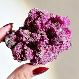 May include: A raw, crystalline mineral specimen featuring a vibrant magenta and deep pink coloration. The cluster has a highly textured, jagged surface with numerous small, irregular facets and dark brown mineral inclusions embedded throughout.