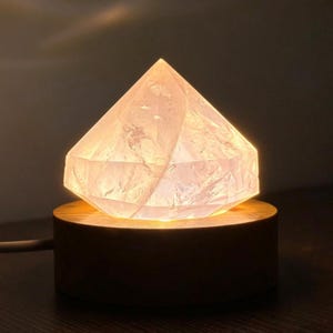 Puede incluir: Una lámpara translúcida en forma de diamante con una superficie facetada que emite un brillo cálido y suave. La estructura similar a un cristal está montada sobre una base circular de madera marrón oscura, con un cable de alimentación lateral.