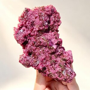 May include: A raw, irregular mineral specimen featuring a vibrant magenta and deep purple crystalline surface. The cluster displays a rough, textured formation with scattered dark brown and metallic inclusions embedded throughout the jagged structure.