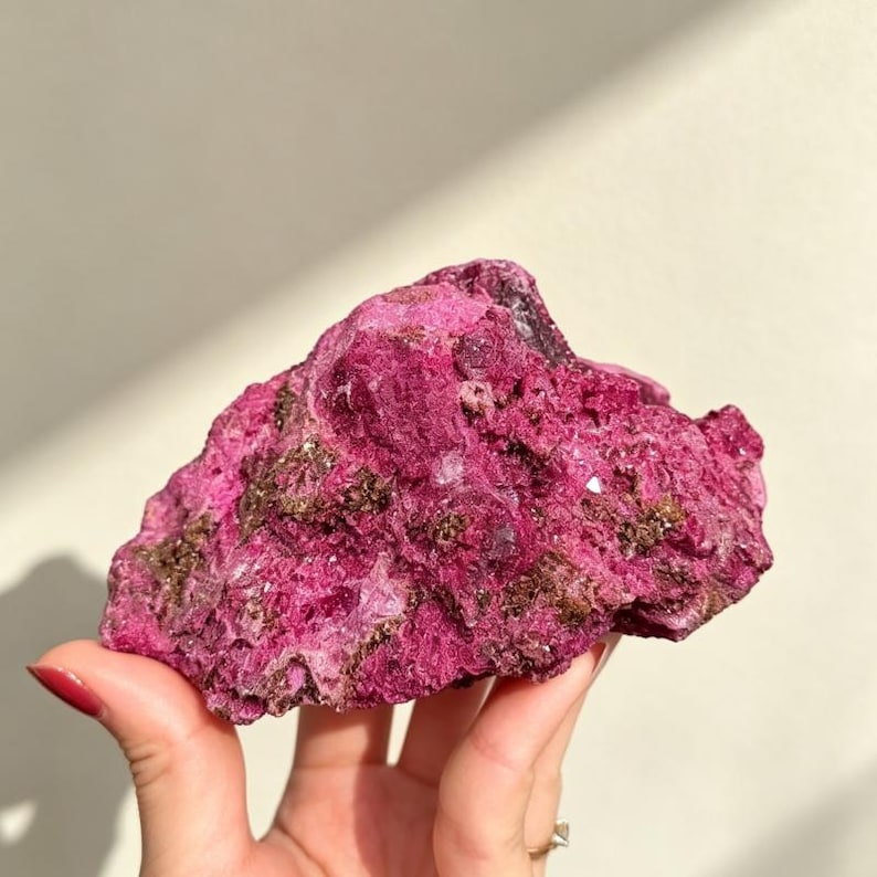 May include: A raw, unpolished mineral specimen featuring a vibrant magenta and deep pink coloration. The surface is highly textured with crystalline formations and small, dark brown metallic inclusions scattered across the jagged, uneven rock structure.