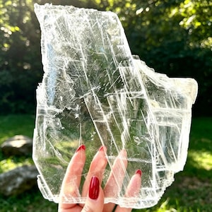 Puede incluir: Una lámina grande, plana y translúcida de cristal de selenita con una apariencia clara similar al vidrio. El espécimen mineral presenta una red compleja de fracturas internas, estrías lineales y bordes afilados y dentados.