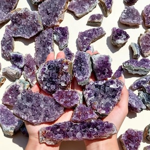 Könnte beinhalten: Eine Sammlung roher Amethyst-Kristallcluster mit tiefvioletten, facettierten Spitzen und gezackten, felsigen Kanten. Die Kristalle variieren in Größe und Form und zeigen natürliche Kristallstrukturen mit durchscheinenden und undurchsichtigen violetten Farbtönen.