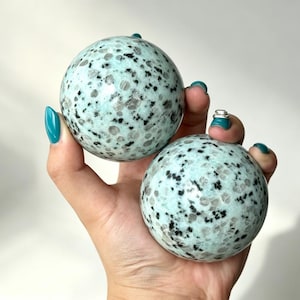 May include: Two polished spherical stones featuring a pale mint green base colour with scattered black and dark grey speckled inclusions throughout the smooth, rounded surfaces of each mineral orb held in a palm.
