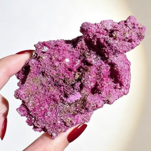 Puede incluir: Un espécimen mineral cristalino en bruto con una vibrante coloración magenta y rosa intenso. La superficie es altamente texturizada con formaciones irregulares y dentadas, además de pequeñas facetas reflectantes que captan la luz.