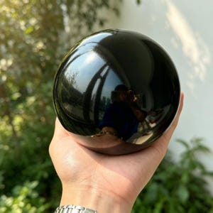 Pode incluir: Uma esfera preta polida e lisa com uma superfície altamente reflexiva. O orbe escuro e brilhante captura a luz e os reflexos do ambiente, exibindo uma cor preta profunda e uniforme em toda a sua forma perfeitamente redonda e sólida.