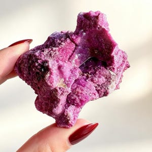 May include: A vibrant, deep magenta-colored mineral specimen with a rough, crystalline texture. The rock features an irregular, jagged shape with a small, hollowed-out cavity in the center, showcasing dense, sparkling clusters of small, pointed crystals.