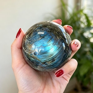 May include: A polished labradorite sphere featuring a dark gray base with prominent iridescent blue and gold flashes across its surface. The stone displays natural internal fractures and a smooth, reflective finish throughout its round shape.