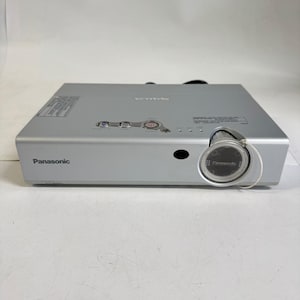 Może przedstawiać: Srebrny projektor cyfrowy marki Panasonic z obiektywem skierowanym do przodu, wyposażonym w osłonę, przyciskiem zasilania oraz przyciskami sterowania na górnej obudowie. Urządzenie ma prostokątny kształt z nadrukowaną nazwą marki Panasonic z przodu.