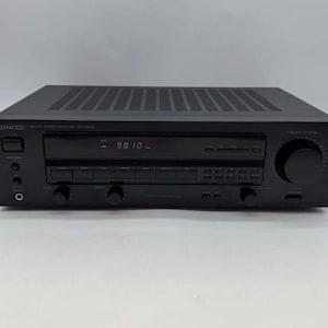 Puede incluir: Un receptor estéreo AM-FM Kenwood modelo KR-A4020 negro con una pantalla digital que muestra 98.10, una perilla de control de volumen circular grande a la derecha, múltiples botones, selectores de entrada y una toma de auriculares en el panel frontal.