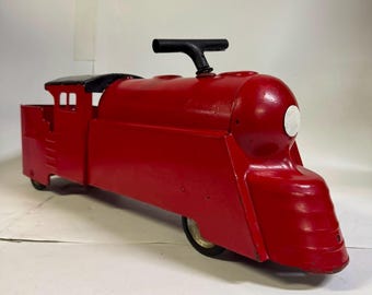 May include: A bright red metal ride-on toy train featuring a streamlined locomotive design, a black T-shaped steering handle on top, a small black roof section over the rear cabin area, and a single white circular headlight detail on the front nose.