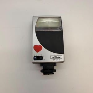 Może przedstawiać: Srebrno-czarna lampa błyskowa Metz 34 CS-2 z prostokątnym panelem świetlnym na górze, czerwoną naklejką w kształcie serca z przodu oraz czujnikiem slave oznaczonym cyframi 2, 1 i 0, wyposażona w czarną podstawę montażową.