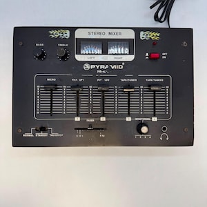 May include: A black Pyramid PR-47 stereo mixer featuring five vertical faders labelled Micro, Pick Up 1, Pick Up 2, Tape/Tuner 1, and Tape/Tuner 2. The unit includes two analogue VU meters, bass and treble knobs, a red power switch, and a black power cord.