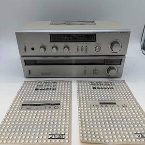 Könnte beinhalten: Ein silberner Sanyo JA 20 Stereo-Vollverstärker und ein Sanyo JT 20 Stereo-Tuner, vertikal gestapelt. Beide Geräte haben silberne Frontplatten mit schwarzen Knöpfen und Reglern. Zwei gedruckte Bedienungsanleitungen liegen vor den Geräten.