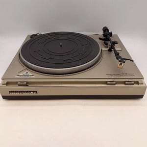 May include: A champagne-colored Marantz TT1200 turntable featuring a black circular platter with a textured rubber mat. The device includes a black tonearm with a gold-toned cartridge, a Marantz logo on the front, and a small warranty sticker.