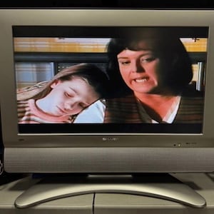 Könnte beinhalten: Ein silberner Sharp-Fernseher mit Breitbildschirm, der eine Szene mit einer dunkelhaarigen Person und einem Kind mit hellbraunem Haar zeigt. Der Fernseher verfügt über ein silbernes Lautsprechergitter unter dem Bildschirm und einen geschwungenen Standfuß.