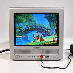Puede incluir: Un televisor Citizen plateado con una pantalla que muestra una escena animada de una persona dibujando cerca de un puente con un perro. El panel frontal cuenta con una rejilla de altavoz, botones de control, un interruptor de encendido y cables RCA rojo y blanco.