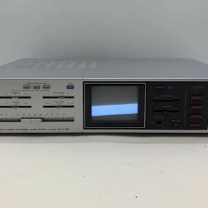 Puede incluir: Un amplificador estéreo de audio y video Akai AV-U8 plateado que cuenta con una pequeña pantalla de monitor integrada, múltiples controles deslizantes horizontales para agudos, graves y balance, un control de volumen, un conector para auriculares.