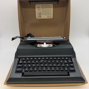 May include: A black electric typewriter with a QWERTY keyboard layout, housed in a tan carrying case. The interior of the case lid features a printed instructional label from Sears, Roebuck and Co. with diagrams for setting up the typewriter.