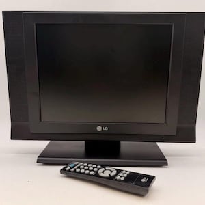 Puede incluir: Un televisor LG negro con pantalla rectangular y altavoces laterales integrados, que presenta un marco grueso y una base de soporte negra a juego. Un mando a distancia negro con botones grises y el logotipo de LG se sitúa frente al televisor.