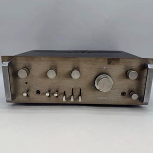 May include: A vintage stereo amplifier with a brushed metal front panel, featuring a large central volume knob, several smaller control dials, and vertical slider switches. The unit has silver-toned handles on both sides and a black metal casing.