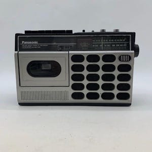 May include: A silver and black Panasonic FM/AM radio cassette recorder featuring a rectangular speaker grille with twenty oval cutouts, a cassette tape compartment, a manual tuning dial, and a frequency display panel with printed text labels.