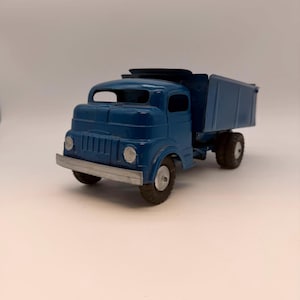 May include: A vintage-style blue metal toy dump truck featuring a silver front bumper, a cab with open windows, and a rear cargo bed. The vehicle is equipped with black rubber tires on silver hubs, showcasing a classic construction design.
