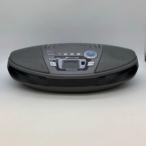 Puede incluir: Un sistema estéreo portátil Panasonic RX-ED50 de color gris oscuro con una pantalla digital central, altavoces integrados a ambos lados, un compartimento para CD de carga superior y varios botones de control, incluido un mando circular.