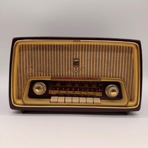 以下が含まれることがあります： ダークマルーン色のケースとクリーム色のフロントパネルを備えたGrundig-Fleetwoodラジオ。スピーカーグリルには垂直の茶色のストライプと中央のエンブレムがあります。2つの金色のノブ、周波数ディスプレイ、6つのボタンが含まれています。