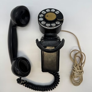May include: A black vintage wall-mounted telephone featuring a circular rotary dial with a brass centre and white lettered inserts. The handset is connected by a black coiled cord, and a separate beige cord extends from the side of the unit.