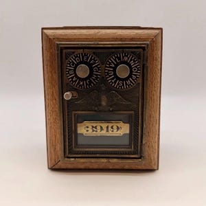 Peut inclure: Une porte de boîte postale ancienne montée dans un cadre en bois. La façade métallique présente deux cadrans de combinaison circulaires avec des lettres, un emblème d'aigle en relief et une fenêtre en verre affichant le nombre 3949 en noir.