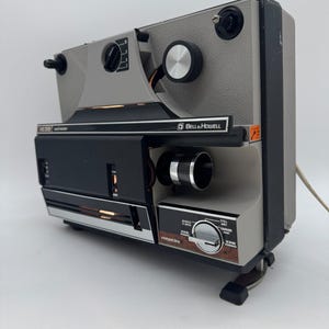 Pode incluir: Um projetor de filmes Bell & Howell 1638 cinza e preto com uma lente proeminente, um botão de controle com configurações para reverso, pausa, avanço e rebobinagem, além de um acabamento texturizado e cabo de alimentação visível.