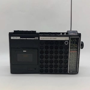 Peut inclure: Un lecteur de cassette et radio Crown Mixing Recorder 475 noir avec une antenne verticale, un lecteur de cassette, une grande grille de haut-parleur circulaire et un cadran de réglage de fréquence vertical sur le côté droit avec des boutons.