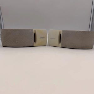 Può includere: Due altoparlanti rettangolari Bose con finitura beige chiaro. Ogni unità presenta una griglia in tessuto testurizzato che copre l'area principale dell'altoparlante e una sezione in plastica liscia sul lato con il logo Bose stampato.