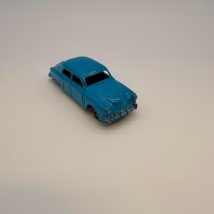 May include: A vintage-style light blue die-cast toy car featuring a saloon body shape, a front grille, and rounded headlights. The paint shows signs of wear and chipping, and the wheels are missing from the chassis of this small metal vehicle.