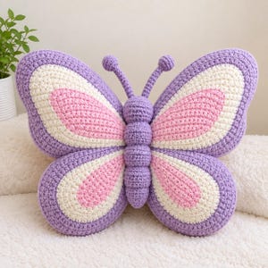 May include: A handcrafted crochet butterfly pillow featuring a central purple body and two antennae. The wings are layered with a purple outer border, a cream-colored middle section, and a soft pink center, creating a symmetrical insect shape.