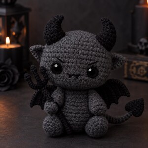 May include: A dark gray crocheted plush toy shaped like a small devil with two black horns, black wings, and a pointed tail. The figure has large black eyes, a small stitched mouth, and holds a black crocheted trident in its right hand.