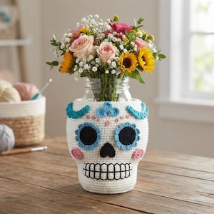 Puede incluir: Un frasco de vidrio transparente con una funda de ganchillo blanca en forma de calavera de azúcar. La funda presenta ojos y nariz bordados en negro, mejillas circulares rosas y detalles florales azules. Un ramo de rosas y girasoles está dentro.