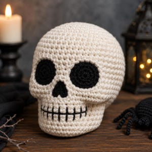 Puede incluir: Una calavera tejida a ganchillo en hilo color crema con grandes cuencas oculares circulares de hilo negro, una nariz triangular negra y una línea negra cosida horizontalmente que representa la boca, con una textura densa y redondeada.